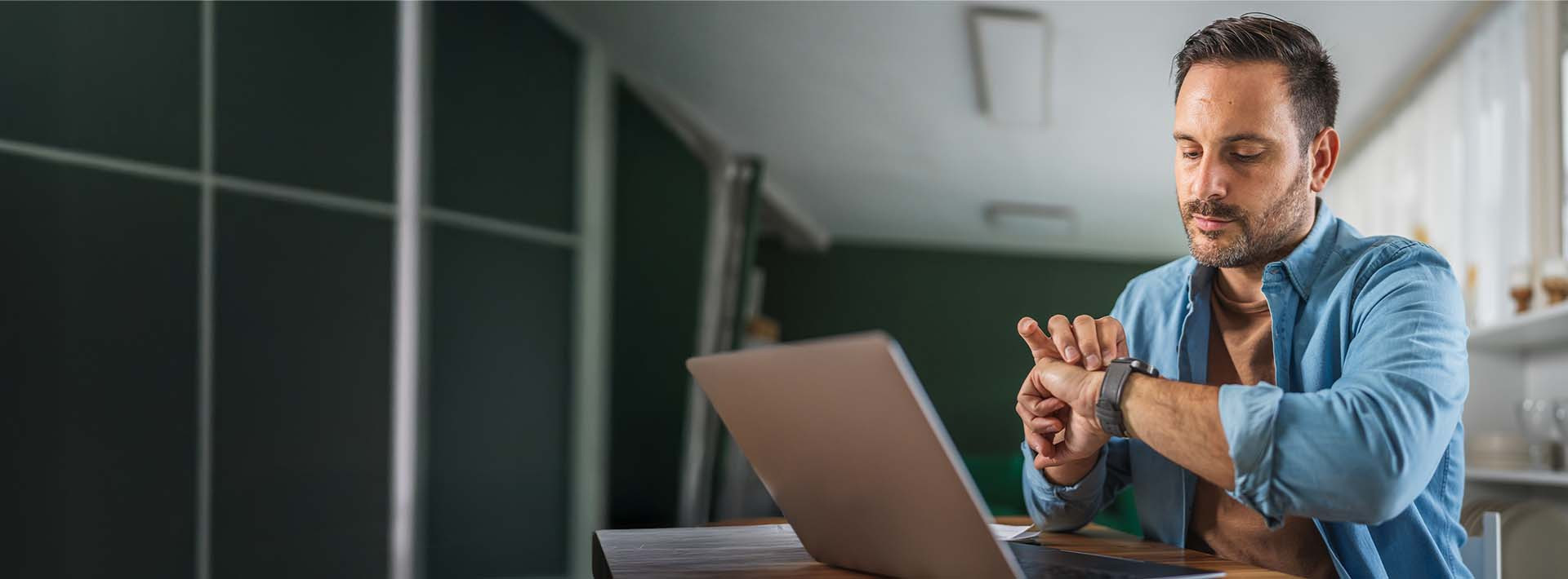 Mężczyzna siedzący przy laptopie, który sprawdza godzinę na zegarku naręcznym, co symbolizuje oczekiwanie na informację.