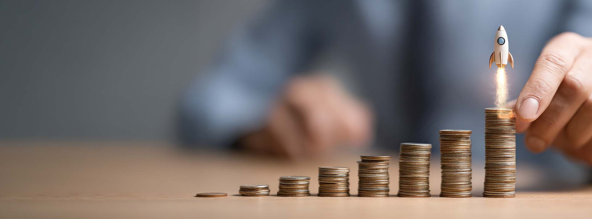 Dłoń układa rosnące stosy monet; nad najwyższym widoczna ikona rakiety symbolizująca wzrost inwestycji lub oszczędności