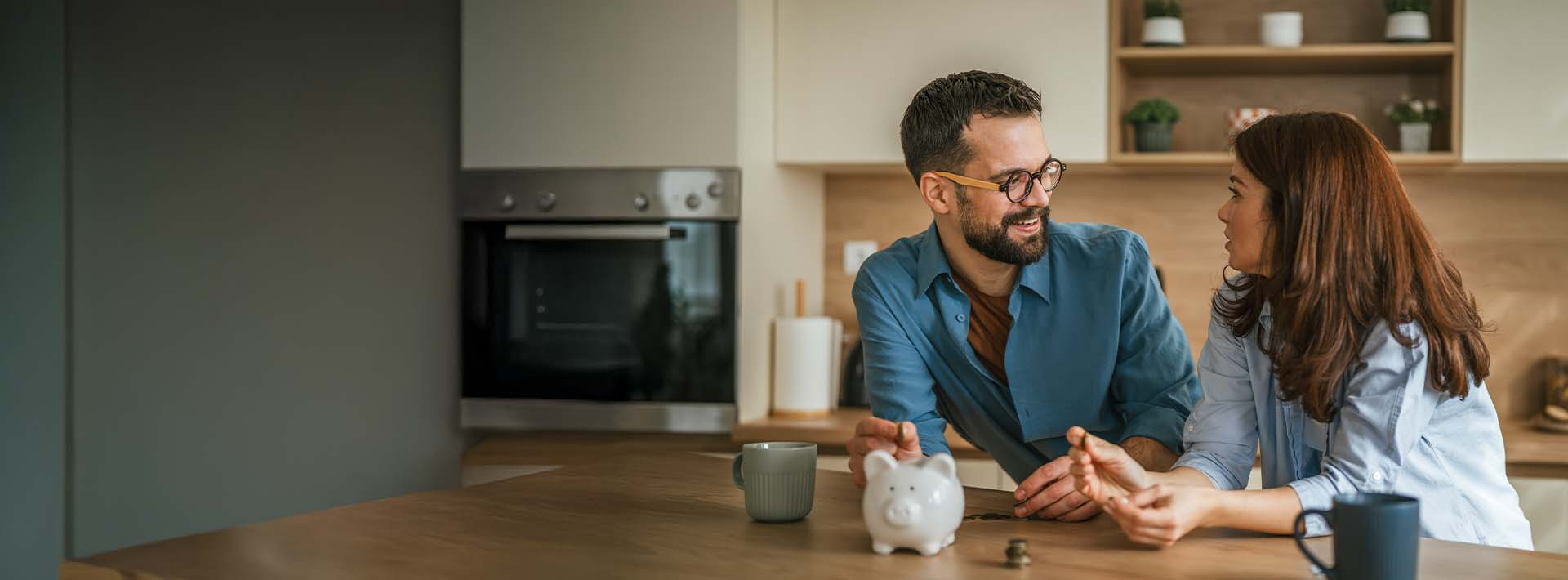 Para siedząca przy stole w kuchni rozmawia i uśmiecha się do siebie, na blacie stoi skarbonka