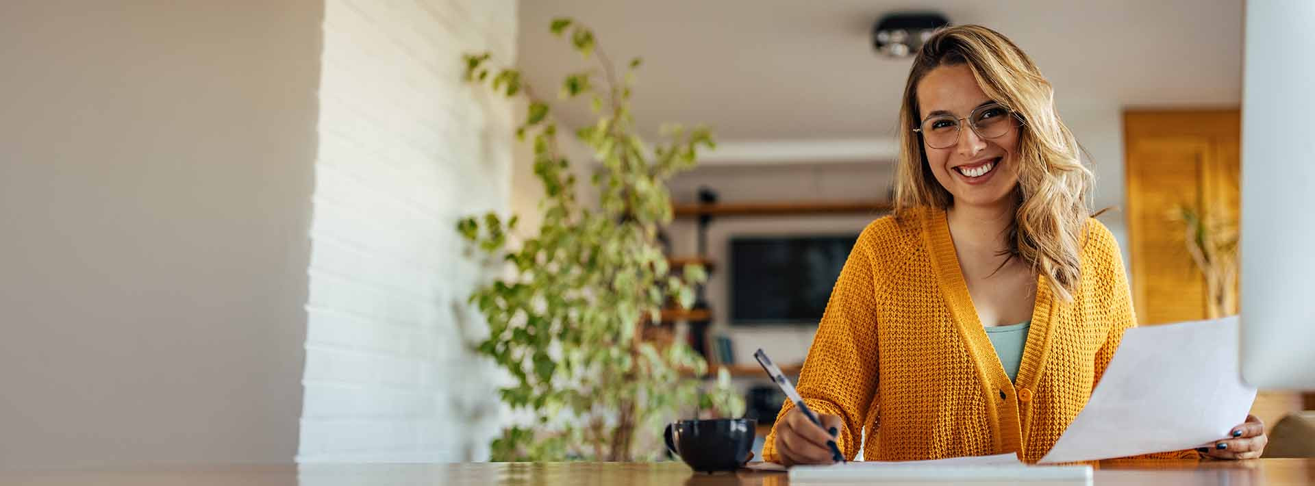 Kobieta w okularach i żółtym swetrze siedzi przy stole, trzyma dokument i długopis; obok stoi filiżanka, pracuje nad notatkami lub rachunkami