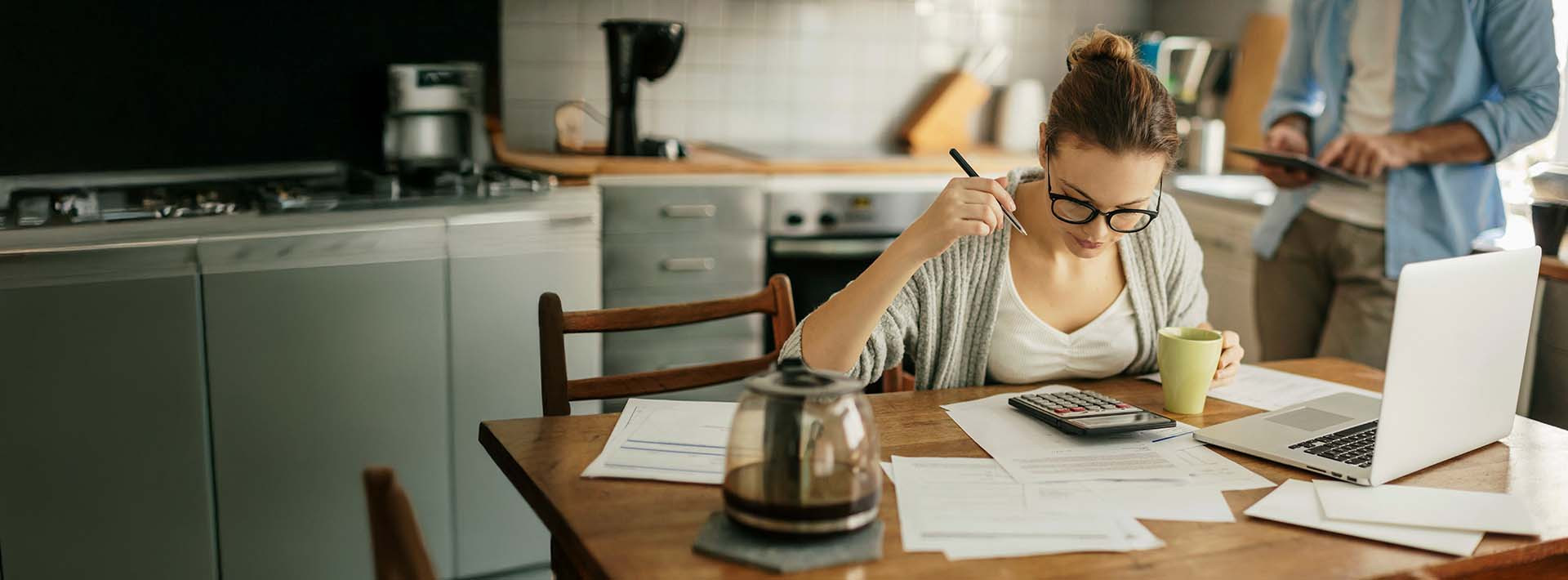 Kobieta siedzi przy stole i przegląda rachunki, korzystając z laptopa i kalkulatora, planując domowe wydatki