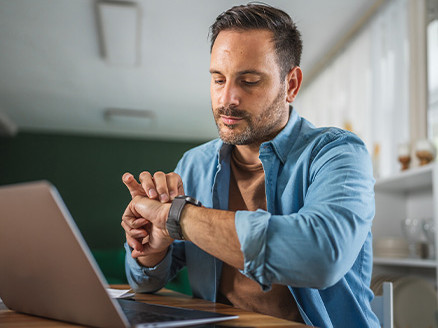 Mężczyzna siedzący przy laptopie, który sprawdza godzinę na zegarku naręcznym, co symbolizuje oczekiwanie na informację.