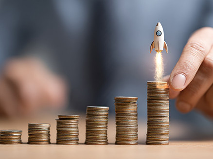 Dłoń układa rosnące stosy monet; nad najwyższym widoczna ikona rakiety symbolizująca wzrost inwestycji lub oszczędności