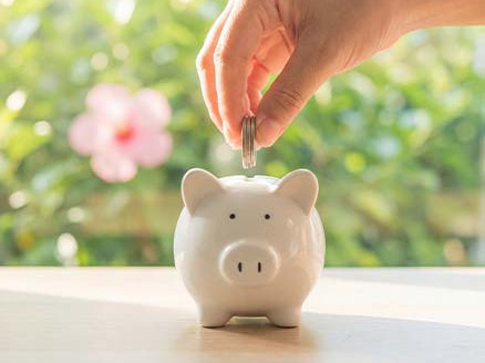 Dłoń wkłada monety do białej skarbonki stojącej na stole, w tle rozmyte zielone rośliny i okno