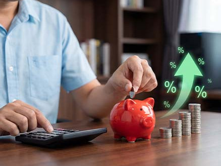 Mężczyzna odkłada monetę do czerwonej skarbonki obok kalkulatora i stosów monet; grafika ze strzałką w górę i symbolami procentów sugeruje wzrost oszczędności lub zysków