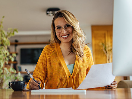 Kobieta w okularach i żółtym swetrze siedzi przy stole, trzyma dokument i długopis; obok stoi filiżanka, pracuje nad notatkami lub rachunkami