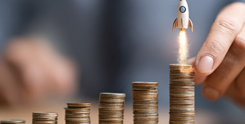 Dłoń układa rosnące stosy monet; nad najwyższym widoczna ikona rakiety symbolizująca wzrost inwestycji lub oszczędności