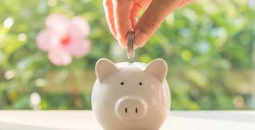 Dłoń wkłada monety do białej skarbonki stojącej na stole, w tle rozmyte zielone rośliny i okno